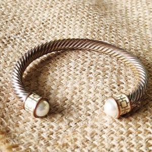 May include: A silver-toned cuff bracelet featuring a twisted cable design. The open ends are capped with cylindrical accents decorated with a cream-coloured geometric pattern and finished with a single round white pearl at each terminal point.