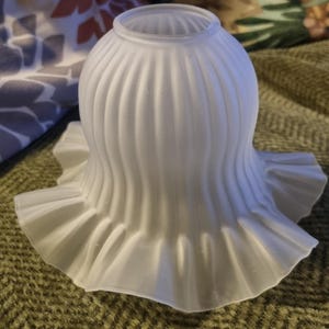 May include: A frosted white glass lamp shade featuring a bell-shaped body with vertical ribbed detailing and a wide, ruffled, scalloped edge at the base. The top opening is circular, designed for attachment to a light fixture socket.