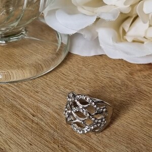 May include: A silver-toned metal ring featuring an intricate, openwork lattice design. The surface of the metal band is embellished with numerous small, clear, sparkling stones set along the interconnected, woven-style metal strands.