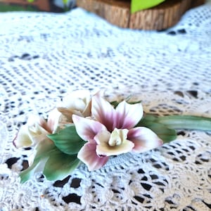 May include: A porcelain orchid figurine featuring delicate petals in shades of white and soft purple, accented by green leaves and a slender stem. The floral arrangement displays intricate details with a smooth finish, capturing the natural form of the orchid.