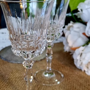 May include: Two clear crystal wine glasses with detailed cut glass patterns. The glasses feature a decorative design on the bowl and stem, and rest on a circular base. The background includes white flowers and foliage, suggesting a formal occasion.