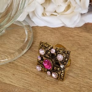 May include: A square-shaped ring with an antique gold-tone finish featuring an intricate filigree metal design. The centre is set with a vibrant fuchsia pink gemstone, surrounded by smaller, pale pink circular crystals embedded in the metal frame.