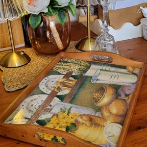 May include: A rectangular wooden serving tray with cutout handles on the short sides. The base features a printed illustration of a kitchen scene with yellow flowers, ceramic plates, a glass pitcher, and bowls of food in warm, earthy tones.