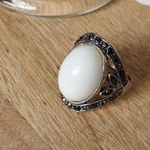 May include: A silver-toned ring featuring a large, smooth, oval-shaped white stone set in the centre. The band and the setting are decorated with small, dark, faceted stones arranged in a leaf-like pattern surrounding the central white stone.
