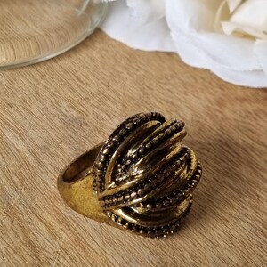 May include: A gold-toned ring featuring a prominent, rounded centrepiece composed of multiple curved, layered bands. Each band is textured with a series of small, raised dots, creating a detailed, dimensional surface across the entire top of the ring.