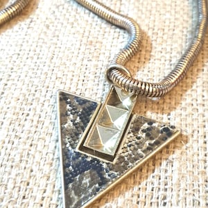May include: A silver-toned metal necklace featuring a triangular pendant with a snakeskin-patterned inlay and a central vertical bar adorned with three raised pyramid studs. The pendant hangs from a flexible, round, silver-toned snake chain.