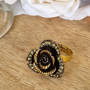 May include: A gold-toned ring featuring a large, dimensional rose-shaped centrepiece. The flower petals are finished in a dark, antiqued black tone with gold highlights, and the outer edges are accented with small, light green sparkling crystals.