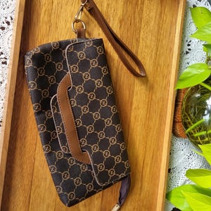 May include: Brown clutch bag featuring a repeating gold-coloured "Z" pattern. It has a brown wrist strap and a contrasting brown flap. The bag is on a wooden tray.