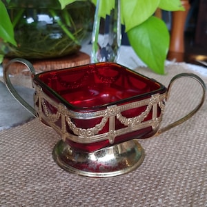 May include: A square, deep red glass bowl set within a gold-toned metal frame featuring ornate swags and vertical pillars. The vessel includes two curved side handles and rests on a circular, pedestal-style base with a metallic finish.
