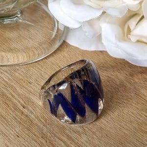 May include: A thick, clear glass ring featuring a swirling internal pattern of deep blue and shimmering copper-coloured flecks. The band has a smooth, rounded exterior surface and a wide profile, showcasing the embedded metallic and blue glass elements.