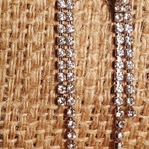 May include: A pair of silver-toned drop earrings. Each earring has a large, round, clear rhinestone at the top, followed by a series of smaller, clear rhinestones in a linear arrangement. The earrings are set against a textured, tan background.