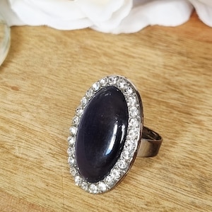 May include: A ring featuring a large, smooth, oval-shaped dark purple cabochon stone set in a silver-toned metal frame. The central stone is surrounded by a border of small, clear, sparkling rhinestones, and the band is a simple silver-toned metal.