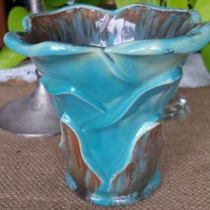 May include: A ceramic vase with a ruffled, flower-like rim, featuring a glossy glaze in shades of turquoise blue and earthy brown. The surface has a textured, organic shape with flowing curves that transition between the blue and brown tones.