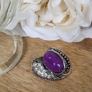 May include: A silver-toned ring featuring a large, smooth, oval-shaped purple cabochon stone set alongside a curved accent piece encrusted with small, clear, sparkling rhinestones, all set within a detailed, textured metal band.