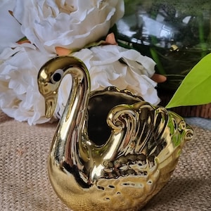 May include: A metallic gold ceramic swan figurine with a glossy finish, featuring a long curved neck, a black and white eye detail, and moulded wing textures on the body. The swan is shaped as a hollow vessel with an open top.