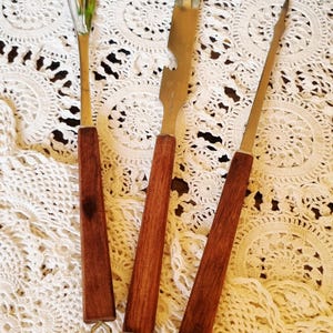May include: A set of three bar tools with dark brown wooden handles and silver-toned metal ends. The set includes a small spoon, a bottle opener with a pointed tip, and a long, thin cocktail pick, each featuring a small metal loop at the base.