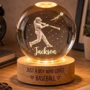 Pode incluir: Uma lâmpada de esfera de vidro transparente com uma gravação a laser 3D de um jogador de basebol a bater e o nome Jackson. A esfera repousa sobre uma base de madeira redonda com o texto JUST A BOY WHO LOVES BASEBALL e corações.