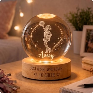May include: A spherical glass night light featuring an etched cheerleader holding pom-poms, the name Avery, and a wooden base engraved with the text JUST A GIRL WHO LOVES TO CHEER and two heart symbols, illuminated by a warm internal light source.
