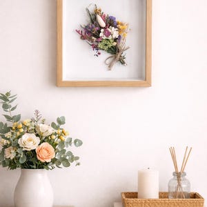 May include: A framed dried flower bouquet with a variety of colors hangs on a white wall. Below, a white vase holds a floral arrangement with peach and white roses. A candle and diffuser sit on a wooden table.