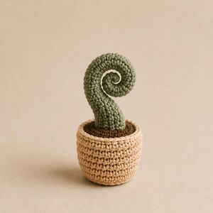 May include: A crocheted plant in a beige pot. The plant has a green, spiral-shaped top, and the pot is filled with brown material. The pot is a light beige color, and the plant is a soft green color.