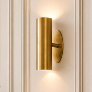 Könnte beinhalten: Zylindrische, goldfarbene Wandleuchte mit kreisförmiger Rückplatte. Die Leuchte strahlt ein warmes Licht aus und wirft einen Schein auf die Wand. Die Leuchte ist an einer weißen Wand mit dekorativen Leisten angebracht. Ein gerahmtes Bild und Grün sind sichtbar.
