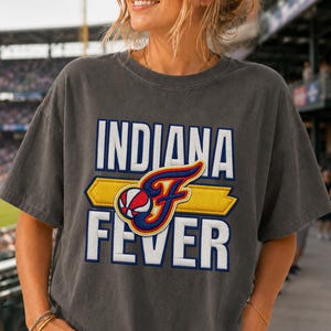 May include: A charcoal gray short-sleeve t-shirt featuring the text INDIANA FEVER in bold white letters. A central graphic displays a stylized letter F in blue, red, and yellow, incorporating a basketball design over a horizontal yellow bar.