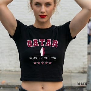A black colored baby tee featuring a Qatar map design with "Soccer Cup '26" and five stars.
