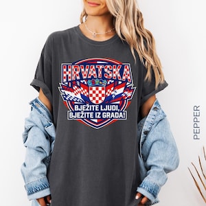 May include: Dark gray t-shirt with a Croatian flag design. The design includes the word "HRVATSKA" in red and white, along with the text "BJEŽITE LJUDI, BJEŽITE IZ GRADA!" in blue and white. The shirt has rolled-up sleeves.
