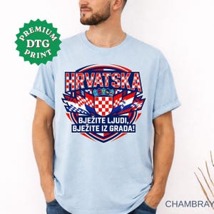 A light blue chambray t-shirt displayed on a bearded man, featuring a large circular graphic with the text "HRVATSKA", the Croatian checkerboard shield, and the slogan "BJEŽITE LJUDI, BJEŽITE IZ GRADA!". A green "PREMIUM DTG PRINT" sticker is visible in the upper left.