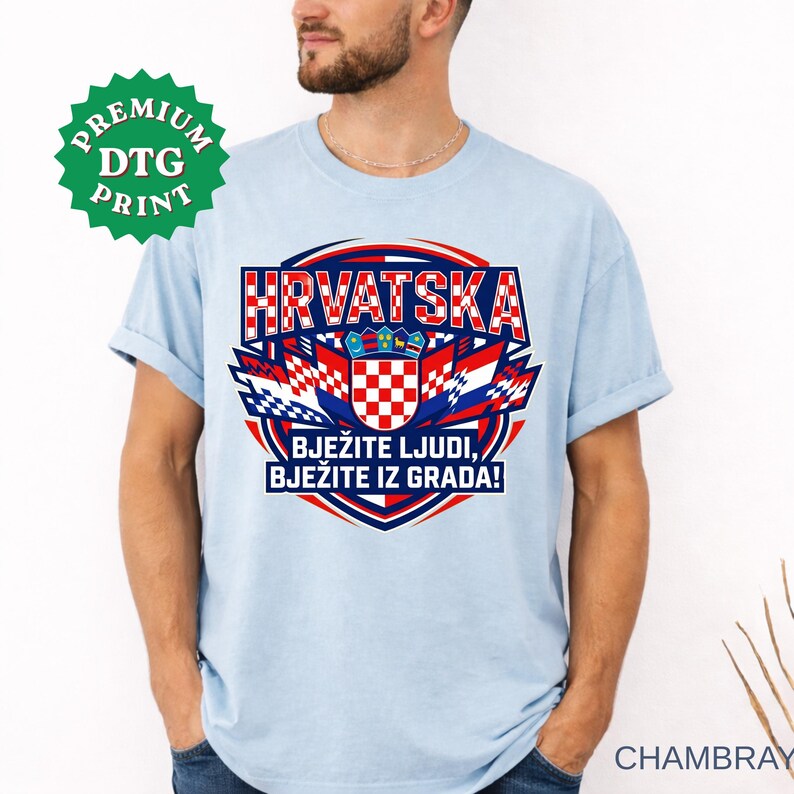 A light blue chambray t-shirt displayed on a bearded man, featuring a large circular graphic with the text "HRVATSKA", the Croatian checkerboard shield, and the slogan "BJEŽITE LJUDI, BJEŽITE IZ GRADA!". A green "PREMIUM DTG PRINT" sticker is visible in the upper left.
