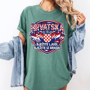 May include: A green t-shirt featuring a Croatian flag design with the word "HRVATSKA" in red and white block letters. The design includes a checkered shield and the text "BJEŽITE LJUDI, BJEŽITE IZ GRADA!"