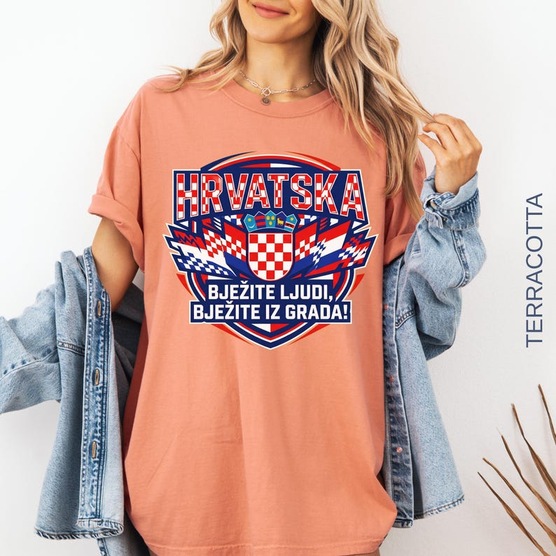 A smiling female model showcasing an oversized terracotta orange t-shirt with the "HRVATSKA" crest design, layered under a light-wash jean jacket.
