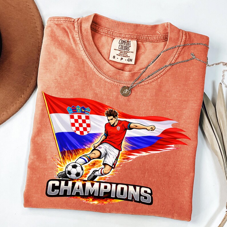 Folded a terracota t-shirt with a graphic design depicting a soccer player kicking a soccer ball, with the word "CHAMPIONS" written below the image.