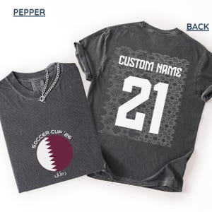 Flat lay of a pepper gray shirt. The front folded view displays the circular maroon and white serrated Qatar flag graphic with curved "SOCCER CUP '26" typography. The flat back view shows a large white square geometric Arabic mosaic pattern under a bold white "CUSTOM NAME" and number 21.