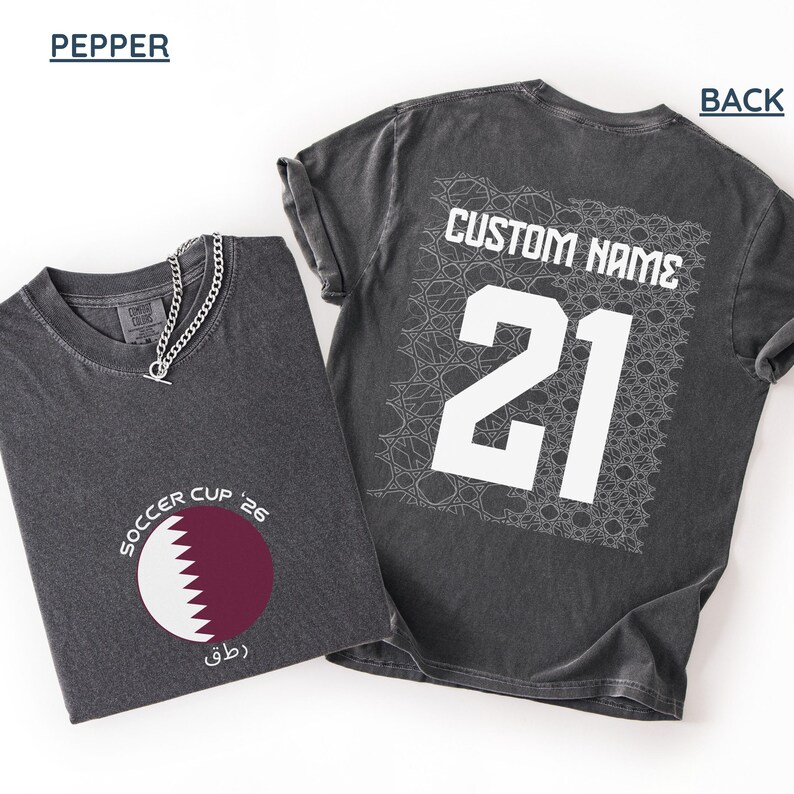 Flat lay of a pepper gray shirt. The front folded view displays the circular maroon and white serrated Qatar flag graphic with curved "SOCCER CUP '26" typography. The flat back view shows a large white square geometric Arabic mosaic pattern under a bold white "CUSTOM NAME" and number 21.
