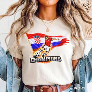 A person wearing a ivory t-shirt with a graphic of a soccer player kicking a ball, along with the text "CHAMPIONS" printed on it.