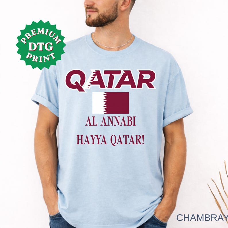 Close-up of a male model wearing a light blue chambray t-shirt featuring the Qatar flag, "QATAR", "AL ANNABI", and "HAYYA QATAR!" text in maroon, with a green "Premium DTG Print" badge overlay in a bright studio setting.