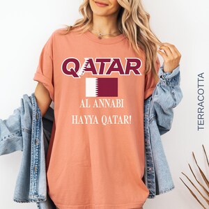 Puede incluir: Camiseta color terracota con la palabra "QATAR" en letras blancas y granates. Debajo, un rect&aacute;ngulo granate con un borde blanco dentado y las palabras "AL ANNABI" y "HAYYA QATAR!" impresas.
