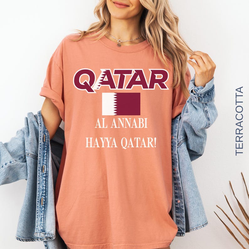 Puede incluir: Camiseta color terracota con la palabra "QATAR" en letras blancas y granates. Debajo, un rect&aacute;ngulo granate con un borde blanco dentado y las palabras "AL ANNABI" y "HAYYA QATAR!" impresas.