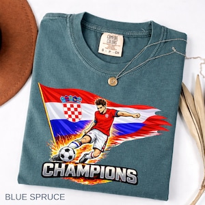 Folded a blue spruce t-shirt with a graphic design depicting a soccer player kicking a soccer ball, with the word "CHAMPIONS" written below the image.