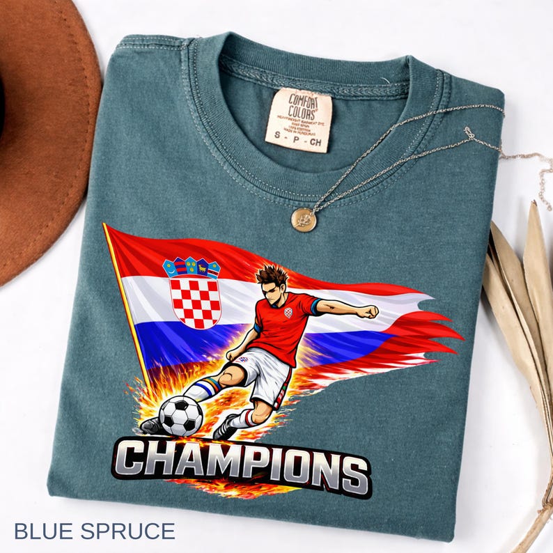 Folded a blue spruce t-shirt with a graphic design depicting a soccer player kicking a soccer ball, with the word "CHAMPIONS" written below the image.