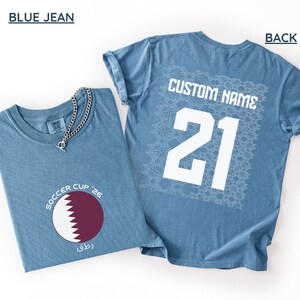 Flat lay of a Blue Jean shirt. The front folded view displays the circular maroon and white serrated Qatar flag graphic with curved "SOCCER CUP '26" typography. The flat back view shows a large white square geometric Arabic mosaic pattern under a bold white "CUSTOM NAME" and number 21.