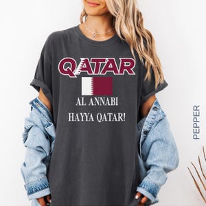 A woman wearing a pepper vintage-style t-shirt with the Qatar soccer design, styled with an open denim jacket and silver jewelry in a bright, modern lifestyle setting.