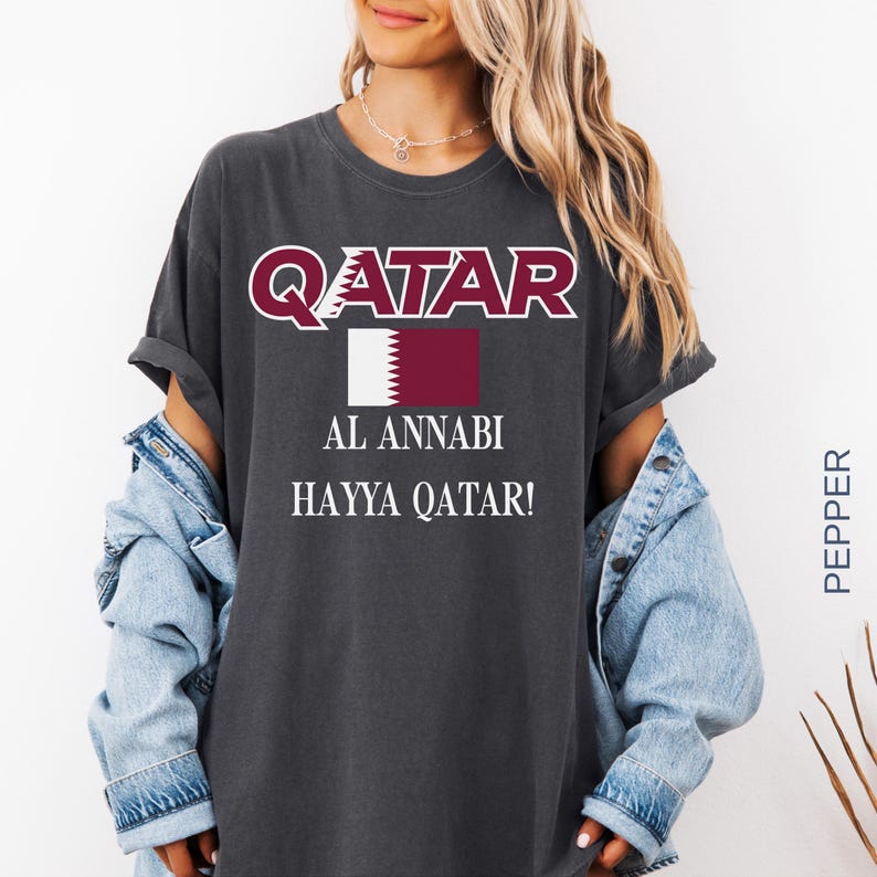 A woman wearing a pepper vintage-style t-shirt with the Qatar soccer design, styled with an open denim jacket and silver jewelry in a bright, modern lifestyle setting.