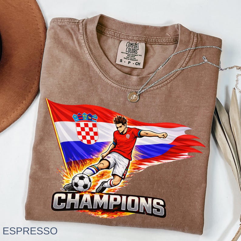 Folded an espresso t-shirt with a graphic design depicting a soccer player kicking a soccer ball, with the word "CHAMPIONS" written below the image.