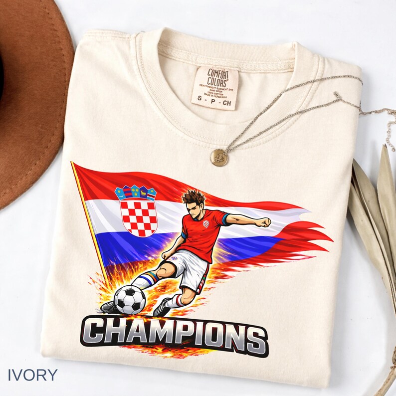 Folded an ivory t-shirt with a graphic design depicting a soccer player kicking a soccer ball, with the word "CHAMPIONS" written below the image.