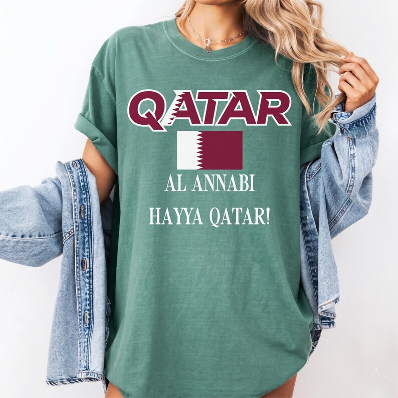 Puede incluir: Camiseta verde con la palabra "QATAR" en granate y blanco, junto con la bandera de Qatar y las frases "AL ANNABI" y "HAYYA QATAR!" en blanco. La camiseta tiene mangas cortas y un corte holgado.