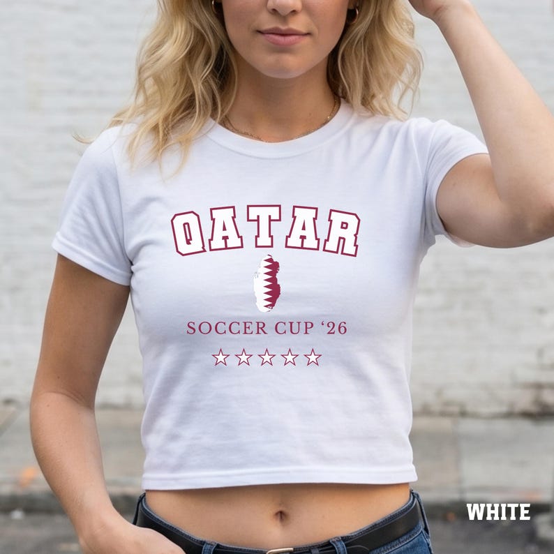 A white colored baby tee featuring a Qatar map design with "Soccer Cup '26" and five stars.