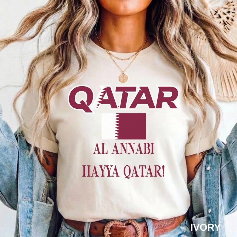 A female model with long wavy blonde hair wearing an ivory t-shirt with the Al Annabi soccer graphic, casually styled with an unbuttoned light wash denim jacket and a brown leather belt.