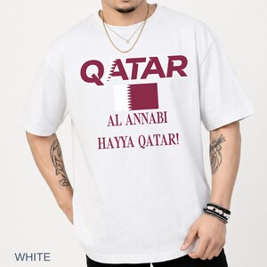 A male model in a crisp white t-shirt showcasing the maroon Qatar football design, styled with layered necklaces and beaded wrist bracelets in a clean, well-lit lifestyle studio.
